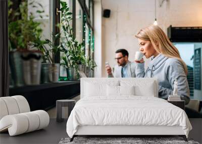 Writing post from cafe. Young woman drinking coffee while working on a laptop with handsome man at the background. Wall mural