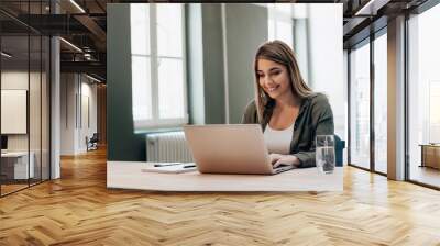 Young female entrepreneur working while sitting at a desk, typing on her laptop computer in a office. Wall mural