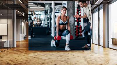 Young muscular woman doing weighted lunge with dumbbells, with personal trainer motivating her. Wall mural