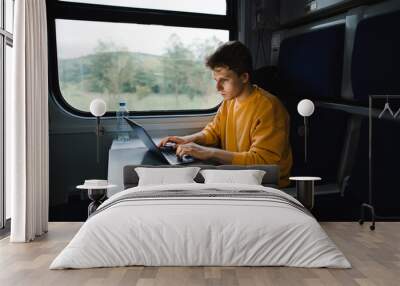 A freelancer man sitting in a train at a table and working on a laptop with a serious face. Freelancer uses laptop on the way by train. Working in travel. Wall mural