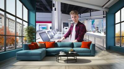 A young man buys a laptop in a technology store. Portrait of a positive young man in the electronics store while choosing a laptop. A look at the camera. Wall mural