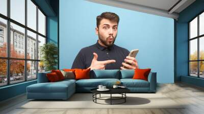 Amazed man in a dark T-shirt and beard, holds a smartphone in his hands, looks emotionally in the camera and shows the index finger on the phone. Emotional man with a smartphone in his hand. Isolated. Wall mural