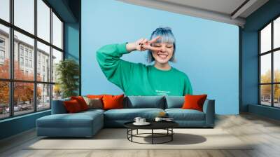 Attractive hipster girl in casual clothes stands on a blue background and shows a gesture of peace winking. Wall mural