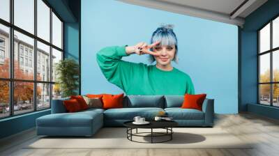 Attractive lady with blue hair poses for the camera with a smile on her face and shows a gesture of peace on a blue background. Wall mural