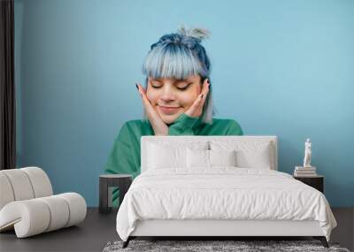 Beautiful lady in casual clothes and colored hair isolated on a blue background, shows a cute face holding her palms to her cheeks and smiling with her eyes closed. Wall mural