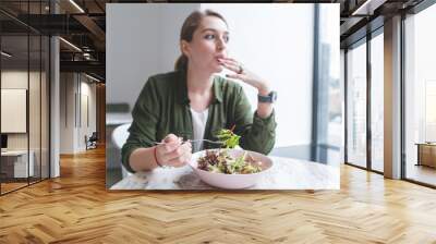 Beautiful young woman eating salad in a restaurant and looking in the window. Focus on a salad plate. Lunch with a healthy meal at the restaurant Wall mural