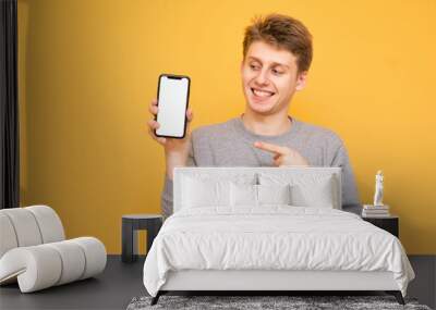 Emotional young man standing on a yellow background holds a smartphone in his hands and looks at the white screen, showing a finger.Smiling guy with a smartphone is isolated on a yellow background. Wall mural