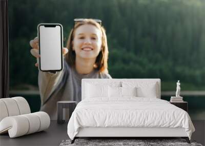 Front view portrait of smiling female traveler showing smartphone blank screen on mountain woods background. Cheerful tourist girl with broad smile demonstrating phone screen at fir forest landscape. Wall mural