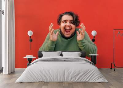 Frustrated young obese bearded guy is shouting and raising his hands up, being angry and emotional, standing in front of a red wall. Wall mural