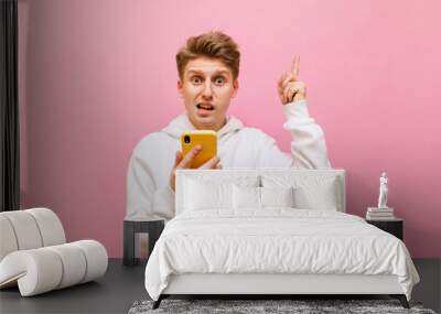 Funny young man with a disgruntled face stands on a pink background with a smartphone in his hands, looks into the camera and shows pointing his finger up at copy space. Isolated. Wall mural