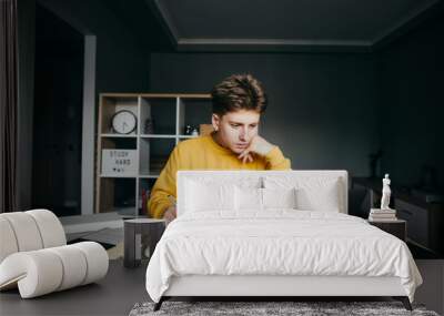 Handsome young man doing homework on desktop at home on room background, looking at laptop screen and writing in notebook. Student studies at home on a remote training in quarantine. Wall mural