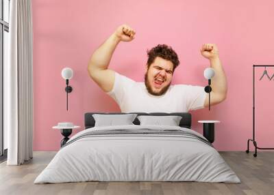 Happy fat young man shouting with joy with his hands raised on a pink background. Joyful overweight guy and white T-shirt, emotionally happy to win. Winning concept. Wall mural