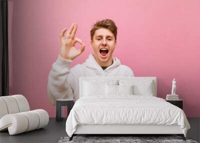 Happy guy in white casual clothes stands on a pink background, shows his thumbs up gesture OK, and looks in camera with a smile on his face.Smiling young man showing thumbs up gesture 