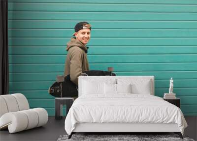 Happy student with a longboard in his hands on a color turquoise background, looking into the camera and smiling. Portrait of a positive skateboarder. Wall mural