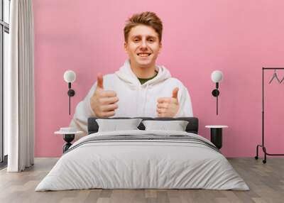 Happy young man in white casual clothes, looks into the camera with a smile on his face and shows thumbs up, isolated on pink background. Cheerful teenager showing gesture like on pink wall background Wall mural