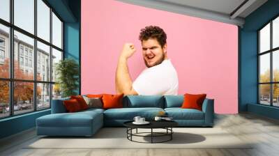 Joyful fat man showing biceps and looking into camera with smile on face isolated on pink background. Portrait of a big boy overweight, happy to win with his arm raised and posing for the camera. Wall mural