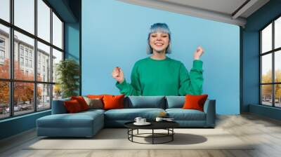 Joyful hipster girl with blue hair stands on a blue background with a happy face raises her hands up and smiles, wears a green sweatshirt Wall mural