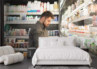 Man wearing a shirt is standing in a supermarket in the milk department and looking at a bottle of milk holds in his hand. Buyer reads the milk label, checks the expiration date. Wall mural