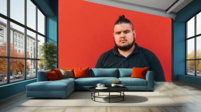 Portrait of a fat young man with a beard on a background of a red wall in a black T-shirt looking at the camera with a serious face. Wall mural