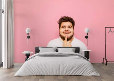 Portrait of a funny happy fat man in white t-shirt on a pink background, looks up at copy space with a smile on his face. A happy overweight guy looks thoughtfully at the empty space above. Wall mural