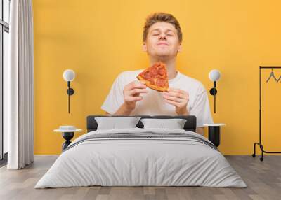 Portrait of a guy in a white T-shirt eats a piece of fresh pizza and gets a pleasure, isolated on a yellow background. Young man is happy to eat a delicious piece of pizza on a yellow background. Wall mural