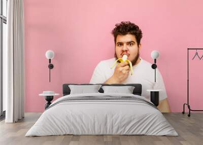 Portrait of funny overweight man with curly hair eating banana on pink background and looking in camera. Fat guy in a white T-shirt bites a banana and looks intently at the camera, isolated. Wall mural