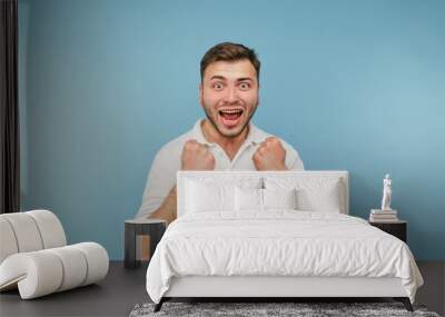 Portrait of joyful adult man rejoices in victory emotionally with raised hands looking at camera and shouting with happiness isolated on blue background Wall mural