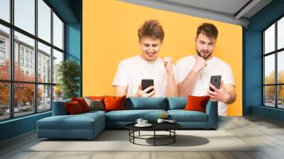 Portrait of two happy male friends standing on a yellow background with smartphones in their hands and rejoicing. Two friends wear a white T-shirt, hold smartphones in their hands and are happy. Wall mural