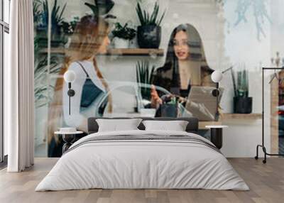 Portrait through cafe window, two happy business women sitting in cozy cafe with laptop and communicate. Two young ladies in formal clothes spend time in a restaurant having a cup of coffee. Wall mural