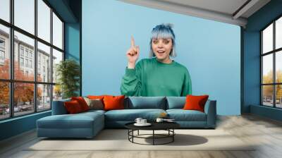 Positive lady with blue hair stands on a blue background with a happy face looking at the camera and showing finger up Wall mural