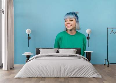 Positive lady with colored hair and in a green sweater stands on a blue background and looks away and laughs. Wall mural