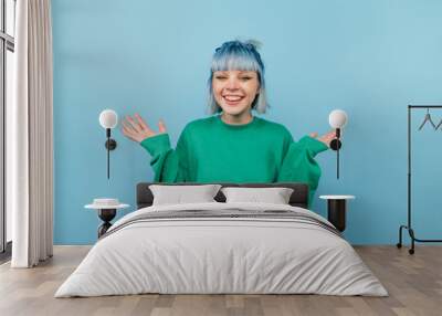 Positive teen girl with blue hair and in a green sweater rejoices with raised hands on a colored background. Wall mural