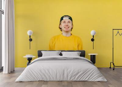 Positive young man in a cap and sweatshirt with a radiant face looking up on a yellow background Wall mural