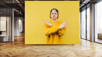 Serious guy in a cap and bright clothes shows a stop gesture and looks focused on the camera, arms crossed on a yellow background Wall mural