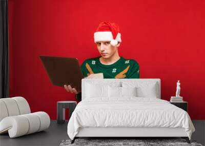 Serious young man in christmas sweater and hat santa stands with laptop on red background and looks intently at camera. Portrait of upset guy with laptop in christmas clothes. Copy space Wall mural