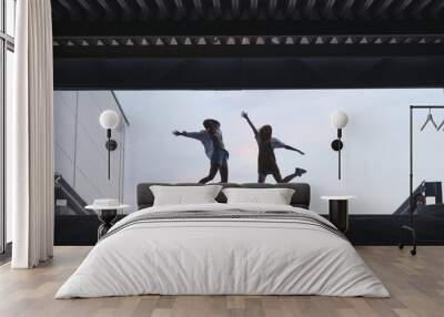 Silhouette of two young girls who jump on. Silhouette of two friends in a jump on the background of the sky near modern architecture. Wall mural