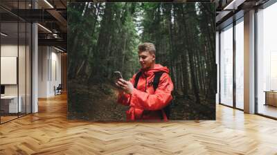 Smiling young man in a red raincoat stands on a forest path in the mountains and uses a smartphone with a positive face. Happy mountain hiker uses smartphone, stands in dark coniferous forest. Wall mural