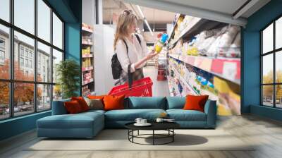 Stylish young girl standing in a grocery supermarket in her arms and reading a label. Female buyer selects products in a supermarket. Wall mural