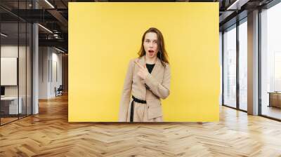 Surprised lady in a beige jacket stands on a yellow background, points her finger to the side and looks at the camera with a shocked face. Wall mural