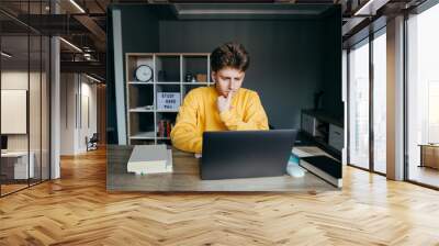 Thoughtful young man in a yellow sweatshirt sits at home at a table in the room and uses a laptop, pensively looking at the screen. Student studying at home using laptop. Quarantine. Wall mural