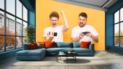 Two young men are competing in a mobile game, standing with smartphones in their hands on a yellow background, one rejoices in victory, the other is tense. Mobile Games Concept. Wall mural