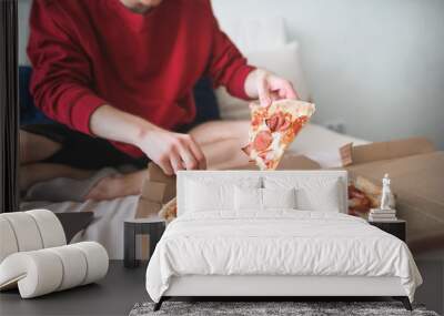 Young man in a red sweatshirt sits at home on a bed and takes an appetizing piece of pizza from the box. Men's hands take a delicious piece of pizza. The teenager is going to eat a piece of pizza Wall mural