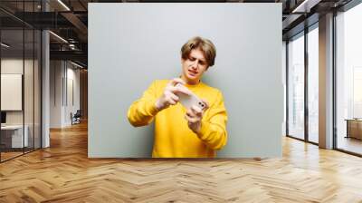 Young man playing online games on a smartphone on a gray background with a focused face, wearing yellow casual clothes. Mobile gaming. Wall mural