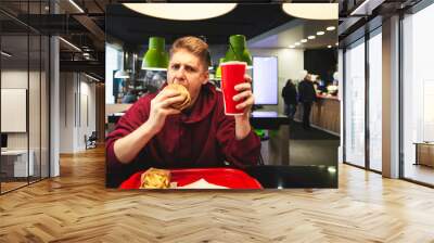 young man sits in a fast food restaurant with a tray of food, cuddles a great appetizing burger and looks at the camera. Portrait of a student dining in a fast food restaurant. Teenager eats a burger. Wall mural