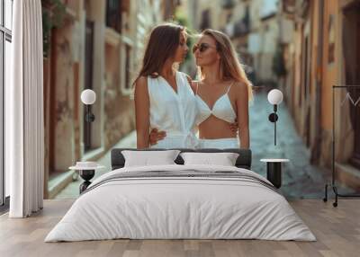 Two fashionable women, dressed in stylish white outfits, share a warm embrace, radiating cheerful summer vibes on a charming cobblestone street that showcases an idyllic urban setting Wall mural