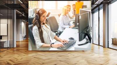 Beautiful friendly female customer support agent with headphones talking with client while using computer in office Wall mural