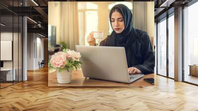 Beautiful young muslim woman wearing abaya drinking coffee and using laptop at home Wall mural