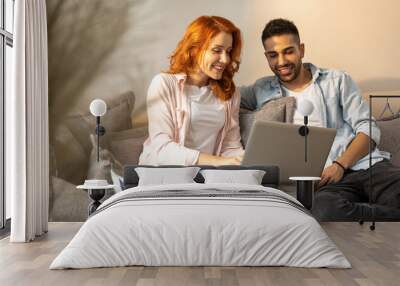 Handsome, young and happy couple watching movie together on a laptop while sitting cuddled up on the sofa at home Wall mural