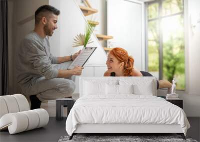 Happy beautiful ginger girl doing exercises in plank position with the assistance of her personal trainer at home. Wall mural