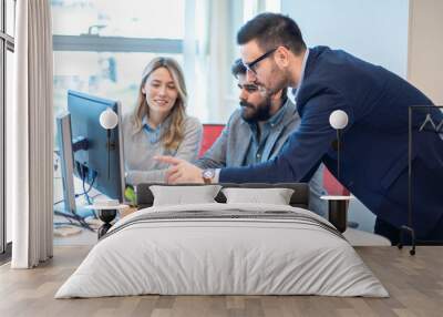 Male executive businessman showing important data on computer screen to his colleague while they are gathered around desk in the office. Wall mural